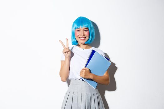 Beautiful asian girl in blue anime wig, showing kawaii peace gesture, holding notebooks, wearing schoolgirl costume for halloween party.