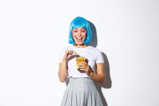 Image of cute asian girl celebrating halloween, eating sweets after trick or treat event, wearing blue short wig and anime schoolgirl costume, standing over white background.