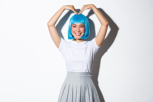 Portrait of cute japanese girl in blue wig showing heart gesture and smiling, standing over white background.