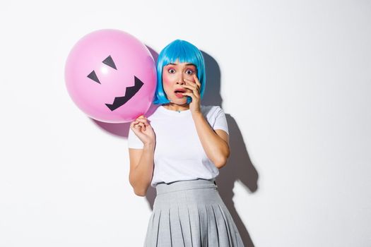 Portrait of scared asian girl in halloween costume and blue wig, gasping and looking ambushed, holding balloon with scary face.