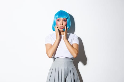 Portrait of surprised asian girl in blue short wig gasping amazed, hear something shocking, standing in halloween costume over white background.