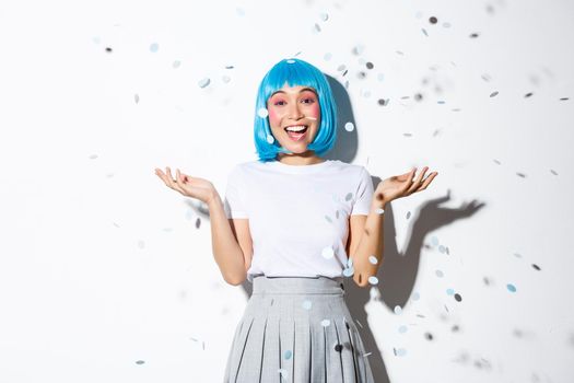 Portrait of happy asian girl in blue wig celebrating halloween, throwing confetti in the air, standing over white background.