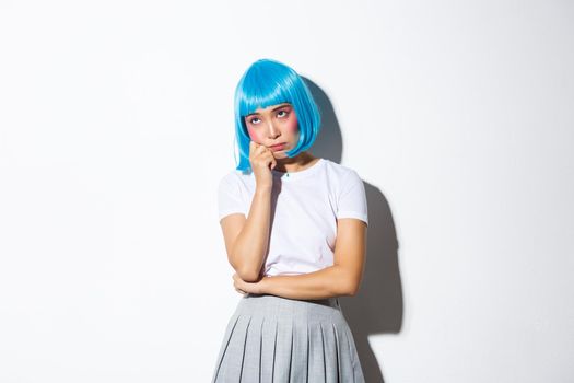 Portrait of moody cute asian girl in blue party wig feeling sad or bored, looking unamused at upper left corner, standing over white background.