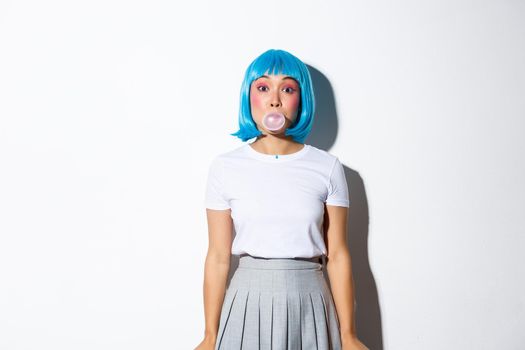 Beautiful asian girl in blue wig and schoolgirl costume, blowing bubble gym with silly face, dressed for halloween party as anime character, standing over white background.