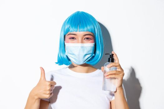 Concept of halloween celebration and coronavirus. Close-up of cute smiling asian girl in blue wig and medical mask showing hand saniziter and thumb-up.
