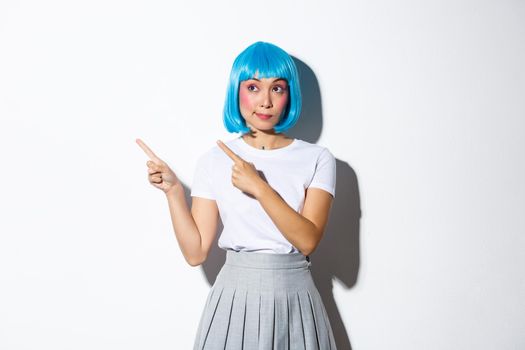 Intrigued silly asian girl with rouge cheeks and kawaii blue wig, looking at upper left corner and pointing fingers at advertisement, showing your logo, standing over white background.