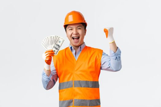 Building sector and industrial workers concept. Happy triumphing asian construction manager, architect in helmet and reflective clothing, fist pump saying yes and showing money, white background.