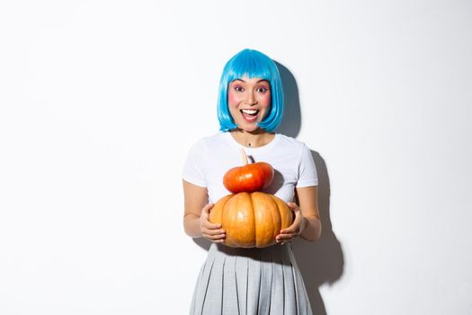 Happy asian girl in blue wig holding two cute pumpkins and smiling at camera, wearing schoolgirl outfit for halloween party.