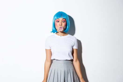 Silly asian girl in blue wig and school uniform, blowing bubble gum with coquettish expression, dressed up for halloween party, standing over white background.