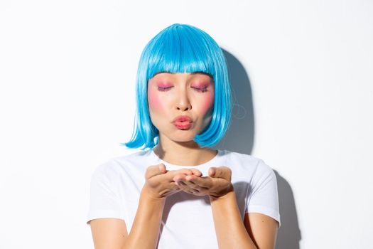 Close-up of silly attractive asian woman with glam makeup and blue wig, blowing air kiss at camera, wearing halloween costume.