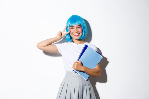 Beautiful asian girl in blue anime wig, showing kawaii peace gesture, holding notebooks, wearing schoolgirl costume for halloween party.