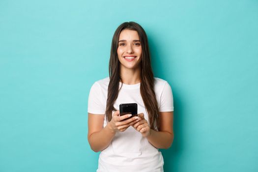 Portrait of attractive smiling woman in white t-shirt, holding smartphone and looking at camera, standing over blue background.