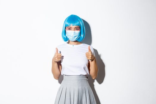 Concept of halloween celebration and covid-19 pandemic. Confident young asian girl showing thumbs-up, wearing medical mask and blue wig.