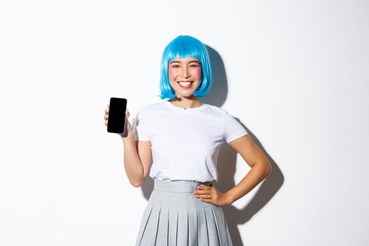 Portrait of cute asian girl dressed as anime character in blue wig, smiling happy and showing smartphone screen, standing over white background.