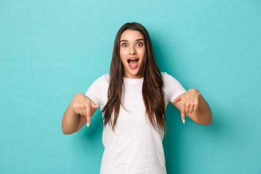 Image of thrilled beautiful girl looking happy and amazed, pointing fingers down, making announcement over blue background.