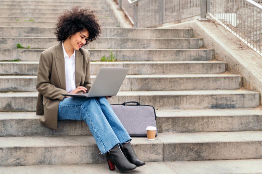 smiling latin woman working with her laptop computer sitting on a city staircase, concept of business and urban lifestyle