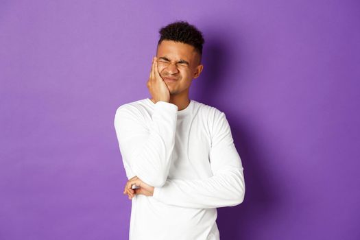 Image of young african-american guy need to see dentist, complaining on toothache, grimacing and touching cheek, being in pain, standing over purple background.