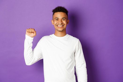 Image of smiling and determined african-american man showing black lifes matter sign, standing united with people of color, showing raised fist and looking at camera, standing over purple background.
