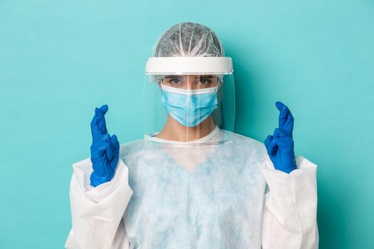 Concept of coronavirus, healthcare and quarantine. Close-up of hopeful female doctor in personal protective equipment, cross fingers and praying, making wish, standing over blue background.