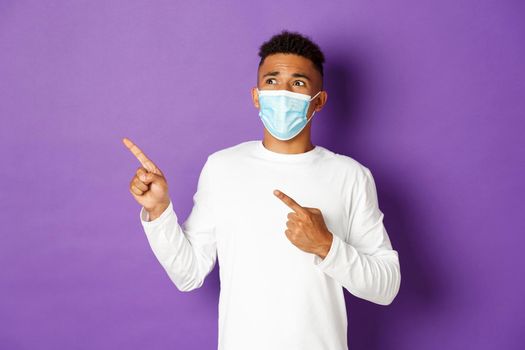 Concept of coronavirus, quarantine and lifestyle. Shocked african-american man, wearing medical mask, pointing and looking nervous at upper left corner, standing over purple background.