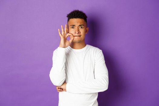 Image of african-american man in white sweatshirt, zipping mouth to make promise, seal lips, keeping secret, standing over purple background.