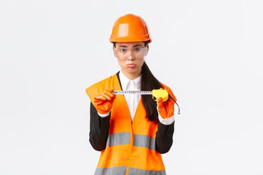 Disappointed gloomy female asian construction engineer, architect showing small size on tape measure, pouting upset with measurements, standing displeased in safety helmet, white background.