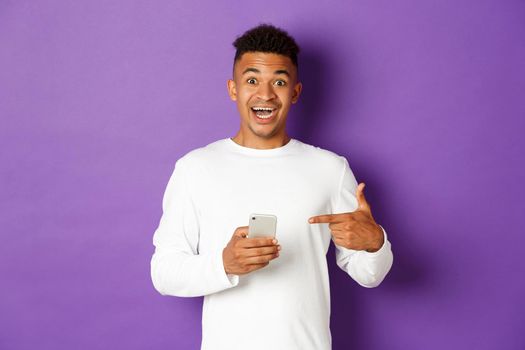 Image of amazed african-american man, pointing finger at mobile phone and smiling excited, recommending app, standing over purple background.