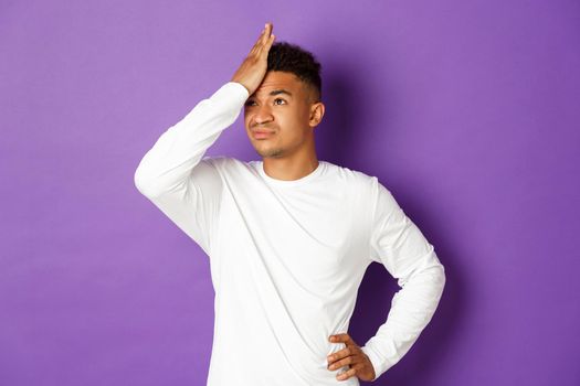 Image of annoyed and bothered african-american male student, slap forehead, making facepalm and rolling eyes from something stupid, standing over purple background.