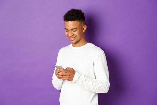Image of young modern african-american man using smartphone, looking at screen and smiling, standing over purple background.
