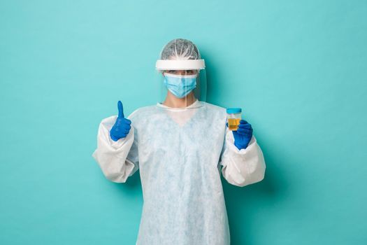 Concept of covid-19, pandemic and health. Satisfied female doctor in personal protective equipment, holding patient urine sample, showing thumbs-up in approval, blue background.