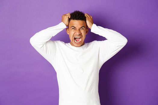 Portrait of tensed and distressed african-american guy losing temper, shouting and ripping hair on head, looking angry up, standing over purple background.