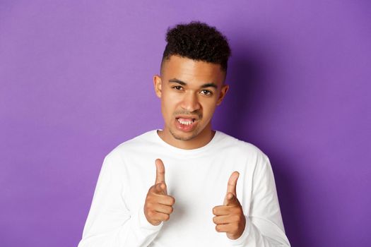 Close-up of cool and sassy african-american guy, pointing fingers at camera, praising good job, congratulate you, standing over purple background.