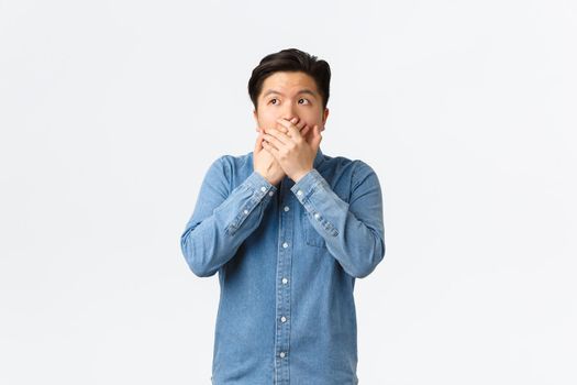 Shocked and speechless asian guy witness something, cover mouth with hands and looking upper left corner, gasping astounded with revelation, hear stunning gossip, white background.