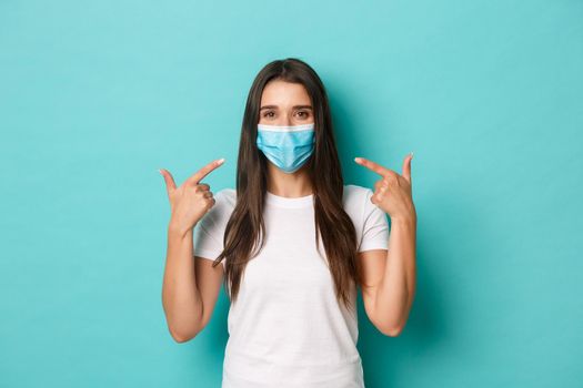 Concept of pandemic, covid-19 and social distancing. Smiling friendly girl in white t-shirt, recommend to wear medical mask during coronavirus, standing over blue background.