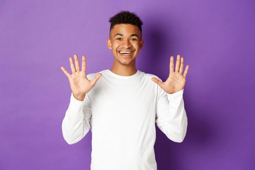 Image of cheerful african-american guy in white sweatshirt, showing number ten and smiling, standing over purple background.