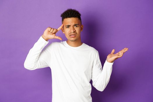 Image of angry african-american man, looking confused, pointing at head and scolding person for stupid action, standing over purple background.