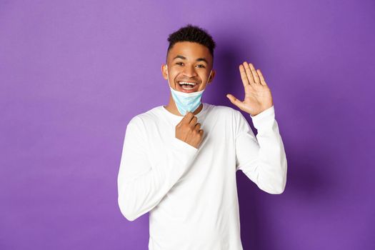 Concept of covid-19, pandemic and social distancing. Image of friendly african-american guy, taking-off medical mask to smile and wave hand, saying hello, greeting someone over purple background.
