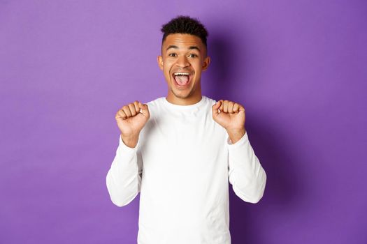 Image of happy and cheerful african-american man in white sweatshirt, jumping from joy, celebrating success or achievement, standing against purple background.