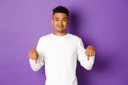 Image of skeptical african-american guy in white sweatshirt, looking disappointed, smirk and pointing fingers down at something unpleasant, standing over purple background.