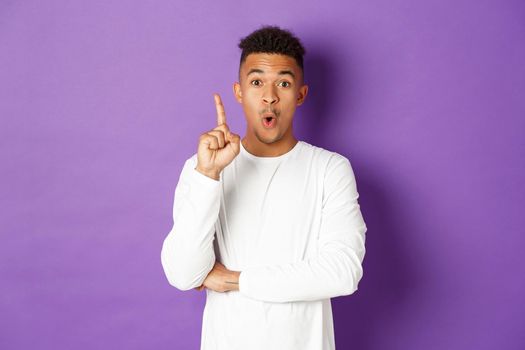 Image of young african-american guy having an idea, raising finger in eureka gesture, suggest plan, think-up solution, standing over purple background.