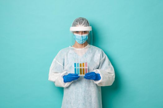 Concept of covid-19, pandemic and health. Image of female lab tech doctor, wearing personal protective equipment, holding test-tubes with vaccine samples, standing over blue background.