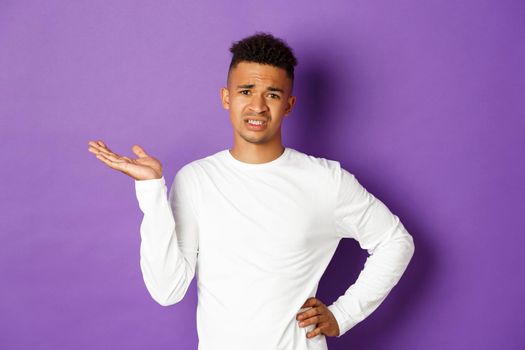 Image of skeptical and unamused african-american man, raising hand up and looking with dismay, standing bothered over purple background.