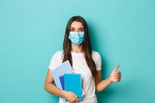 Concept of coronavirus, health and social distancing. Portrait of female student attend classes in medical mask, showing thumbs-up in approval, holding notebooks, standing over blue background.