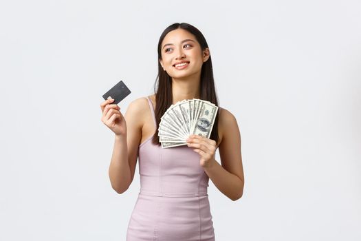 Luxury women, party and holidays concept. Dreamy and excited smiling rich asian woman in evening dress, looking away imaging what buy on cash, holding credit card and money.