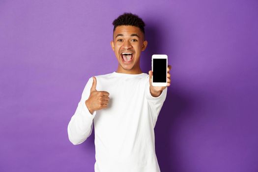 Image of african-american guy in white sweatshirt, smiling and showing thumbs-up in approval, showing mobile phone screen, standing over purple background.