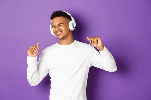 Portrait of handsome modern african-american guy, listening music in headphones and dancing carefree, standing over purple background.