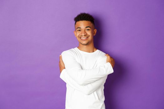 Cheerful african-american man in white sweatshirt, embracing himself and smiling, standing happy over purple background.