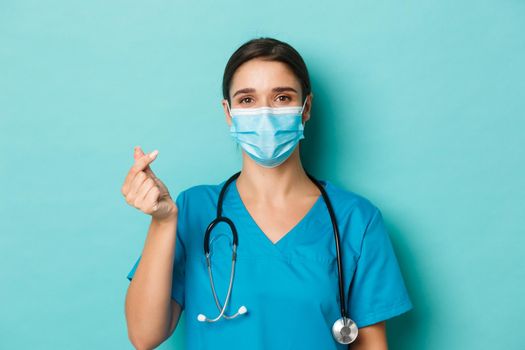 Concept of covid-19 and quarantine concept. Cheerful beautiful female doctor in scrubs and medical mask, showing heart sign, standing over blue background.