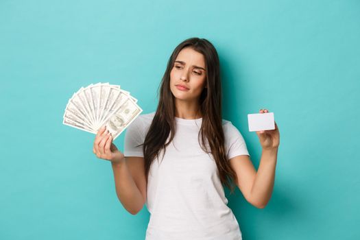 Image of thoughtful young woman making choice between cash and credit card, standing over blue background.