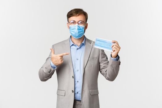 Concept of work, covid-19 and social distancing. Image of male employer in gray suit, encourage using medical mask in office, standing over white background.